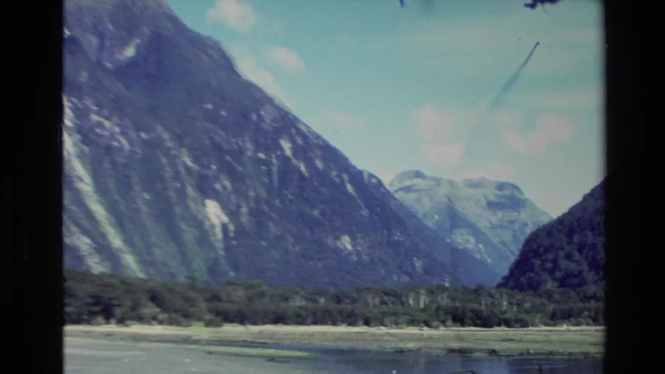 1982 New Zealand 8mm tourist footage showing forested mountain slopes, distant pale peaks, and a low shoreline across water.