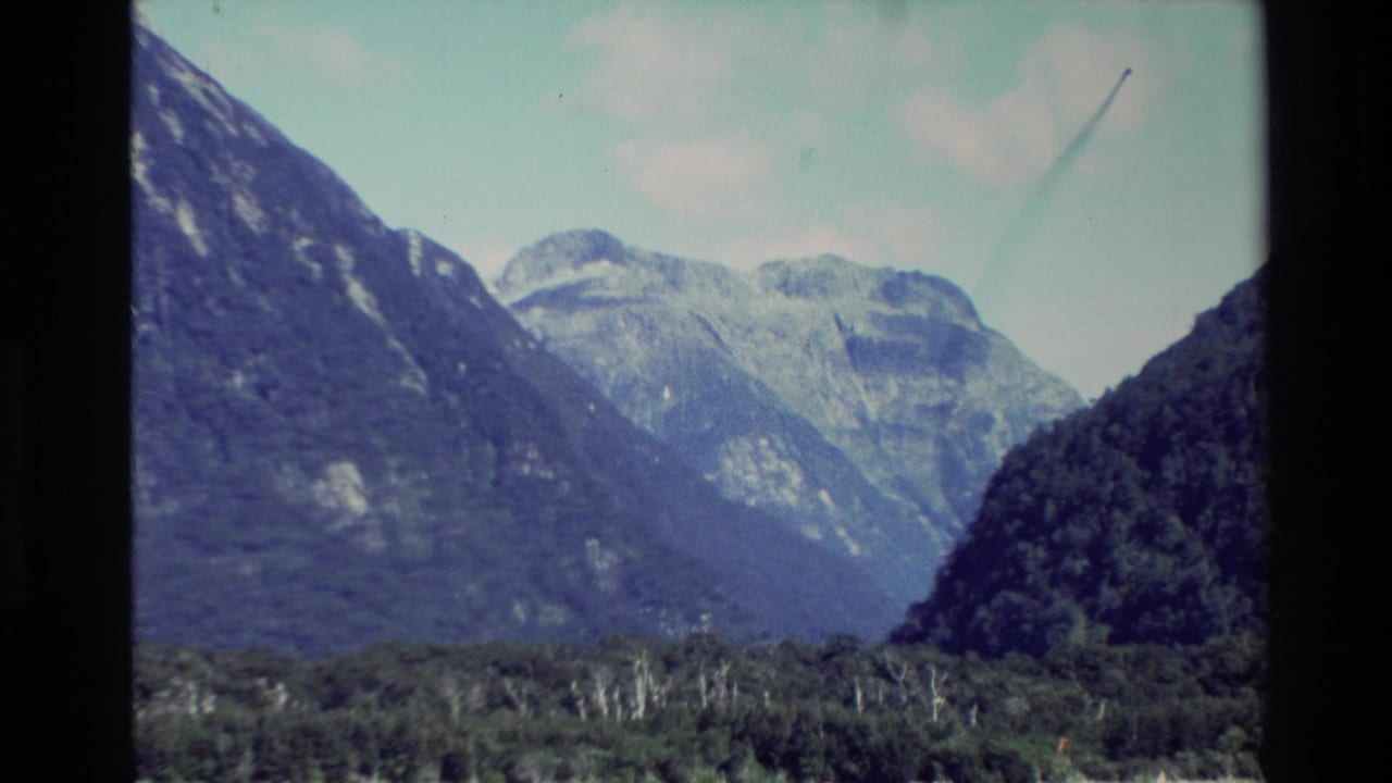 New Zealand mountain scenery on 8mm film, with steep dark slopes, a distant rocky summit, forest below, and visible frame wear.