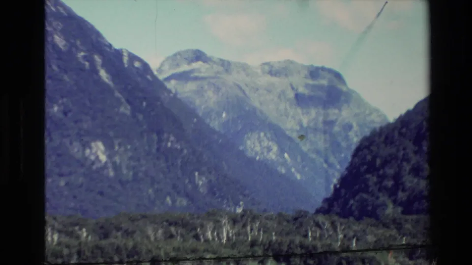Soft 8mm view of New Zealand mountains, with pale rock faces rising beyond dark wooded hills and visible home-movie grain.