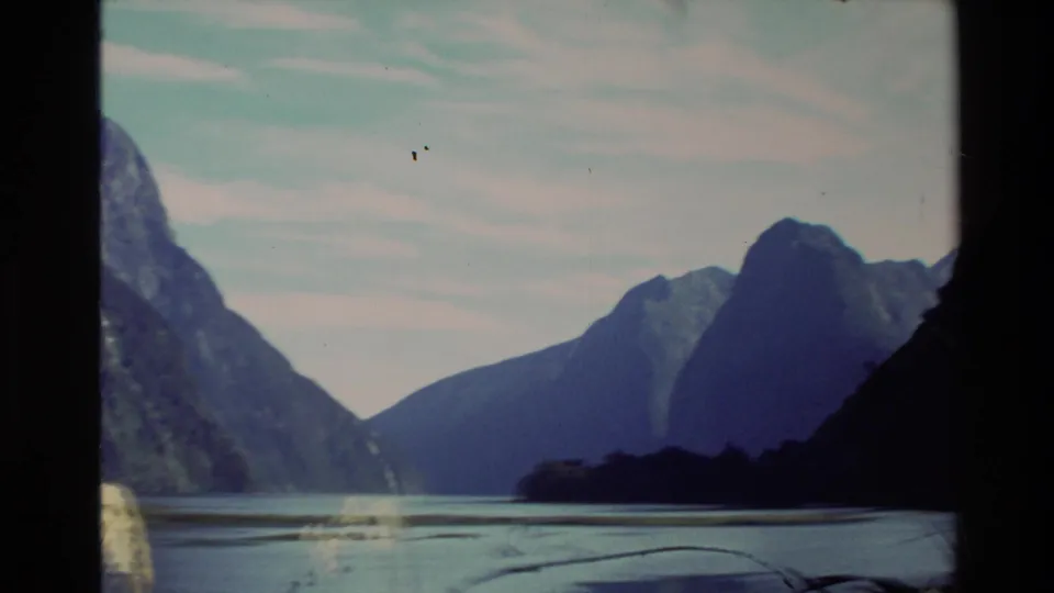 1982 New Zealand home-movie frame showing a calm lake or inlet between dark mountain slopes, with distant peaks and pale clouds.