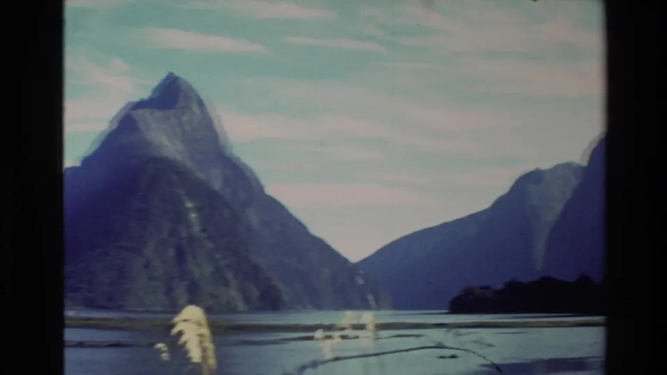1982 New Zealand 8mm frame showing a steep mountain beside calm water, with shoreline reeds and pale sky visible.
