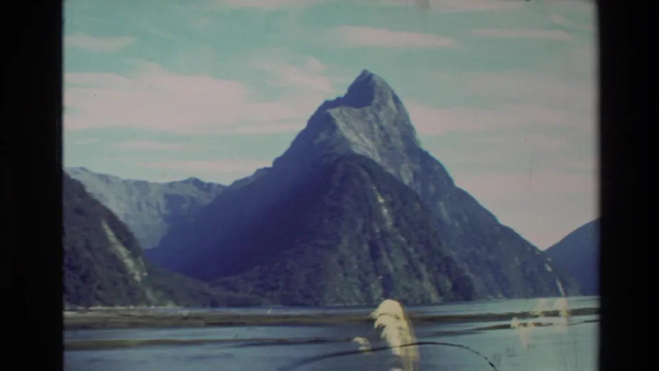 1982 New Zealand 8mm frame with a prominent mountain peak, calm water, foreground reeds, and soft film edges.