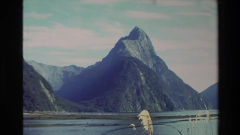 1982 New Zealand 8mm frame showing a mountain across calm water, with distant slopes and reeds near the shore.