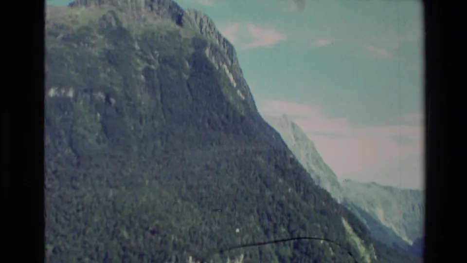 1982 New Zealand 8mm frame showing a closer forested mountainside, with water below and pale sky along the edge.