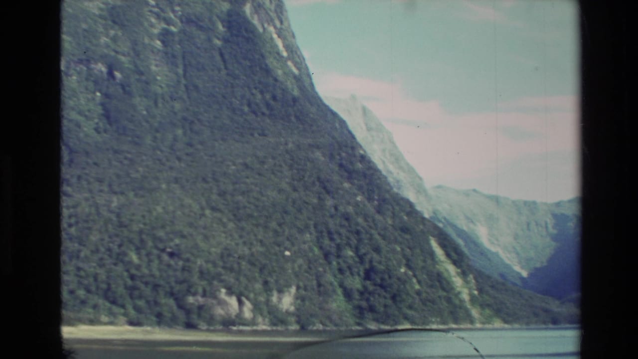 Steep forested mountain slopes rise from calm water under a pale sky.