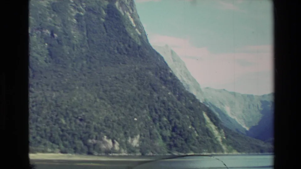 1982 New Zealand 8mm frame showing forested fjord mountains and calm water with soft film grain and dark frame edges.