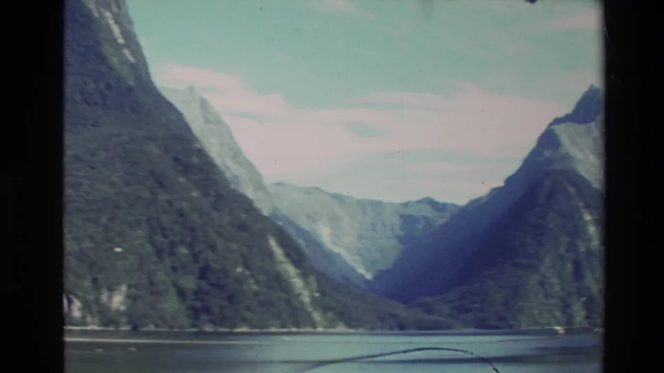 1982 New Zealand home-movie frame looking across still water toward steep green mountains and a shadowed valley.
