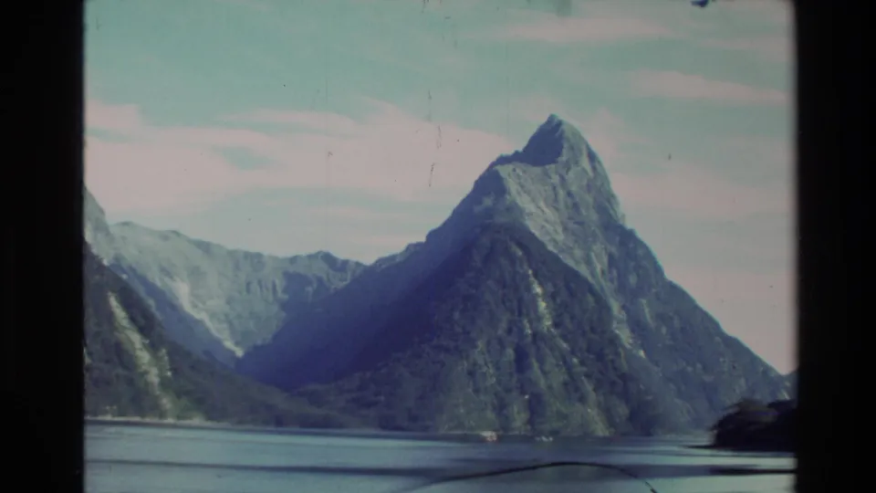1982 New Zealand 8mm frame featuring a sharp pyramid-like mountain peak above calm fjord water and hazy ridgelines.