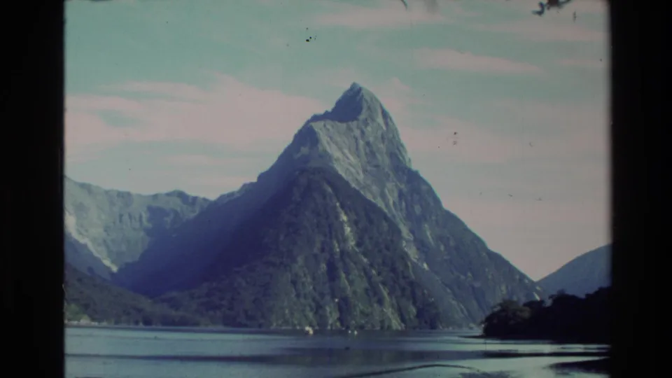1982 New Zealand home-movie frame with a prominent pointed peak, calm water, dark shoreline, and visible Super 8 texture.