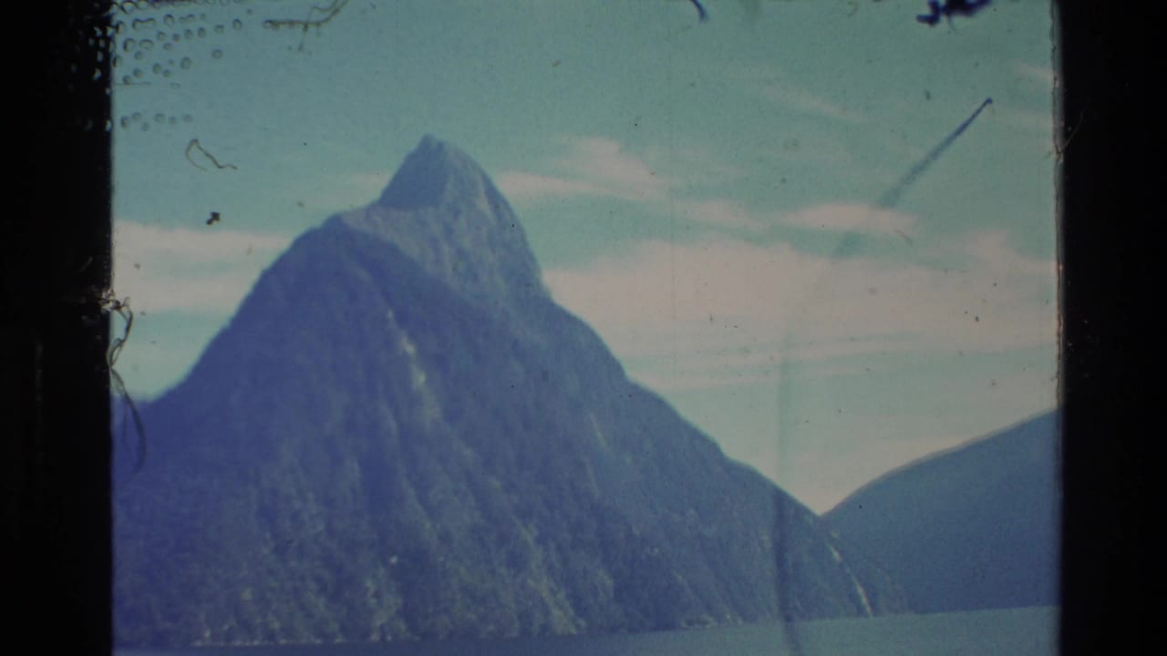 Tall mountain peak rising above calm lake water beneath a pale cloudy sky.