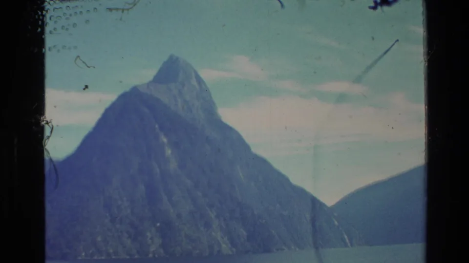 1982 New Zealand 8mm frame showing a steep mountain peak across blue-green lake water under a lightly clouded sky.
