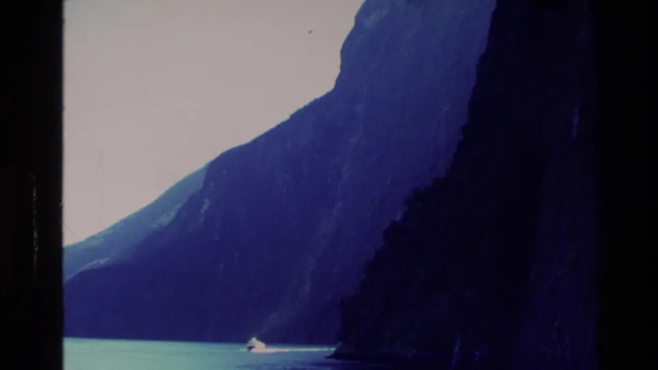 1982 New Zealand 8mm frame showing a steep dark cliff beside blue-green lake water, with a small bright wake near the shoreline.