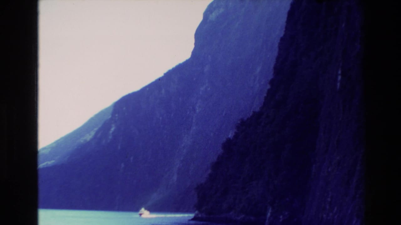1982 New Zealand 8mm frame showing a small boat moving through turquoise water beneath steep, shadowed mountain cliffs.