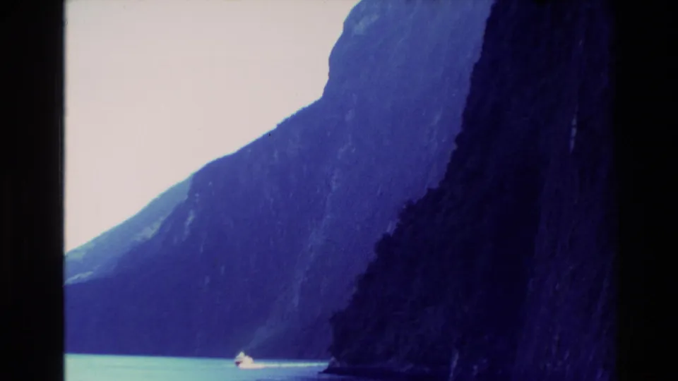 1982 New Zealand 8mm frame showing a small boat moving through turquoise water beneath steep, shadowed mountain cliffs.