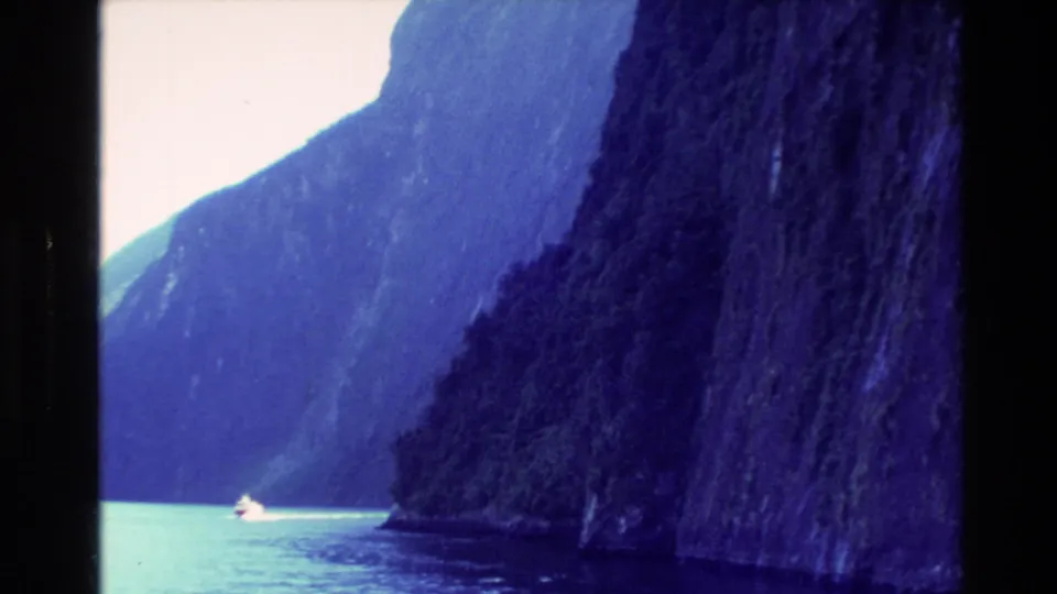 1982 New Zealand travel footage frame with a small boat near a dark cliff and layered blue mountain walls across the water.