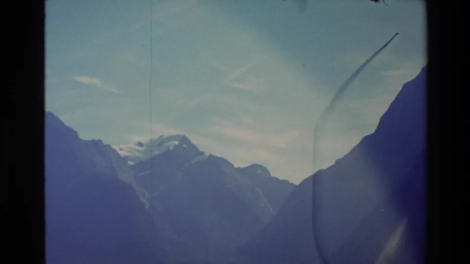 1982 New Zealand 8mm frame looking toward snow-capped peaks between dark mountain slopes under a pale sky.
