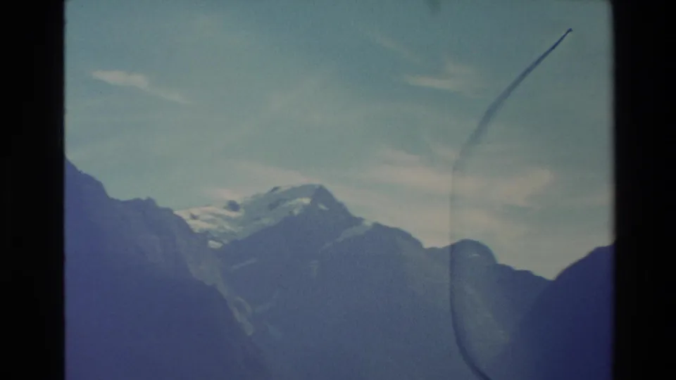 1982 New Zealand home-movie frame of a distant snow-capped peak framed by blue mountain silhouettes and visible film flare.