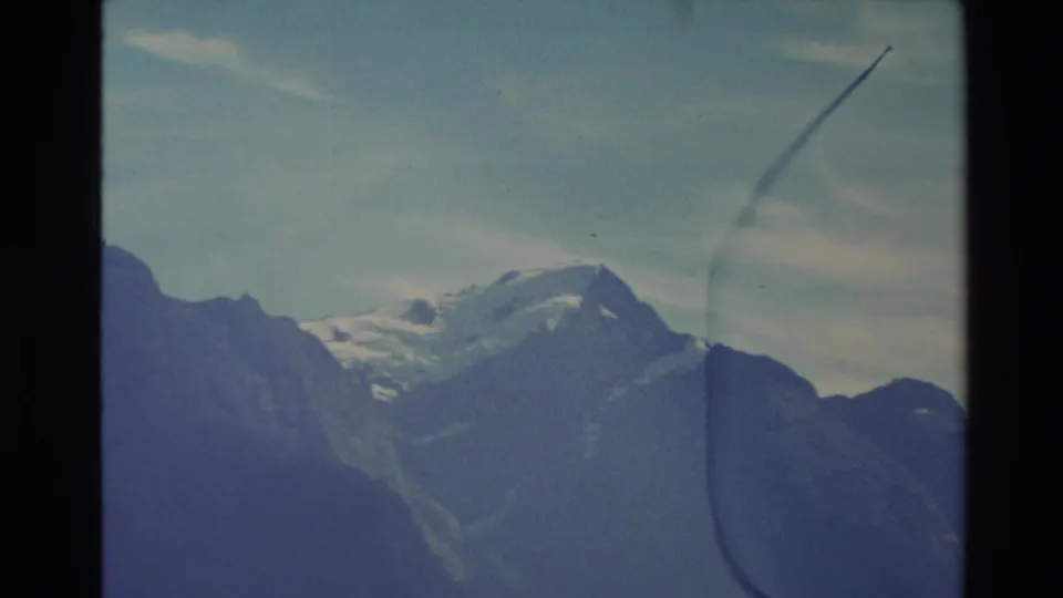 1982 New Zealand 8mm frame showing snow on distant mountain peaks above shaded alpine slopes and a clear sky.