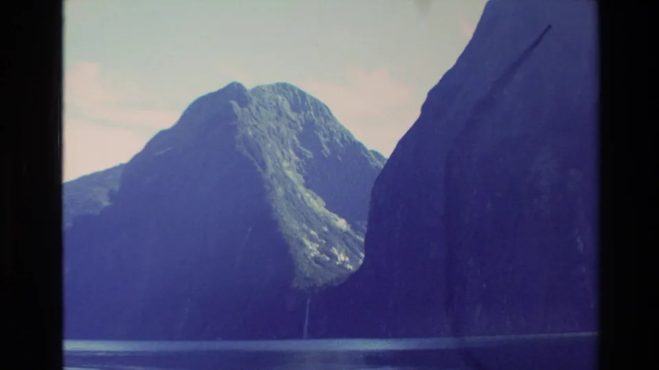 1982 New Zealand 8mm frame showing steep alpine walls, calm lake water, and a thin waterfall in a valley.