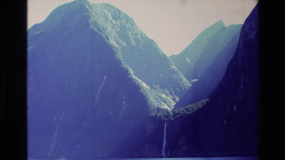1982 New Zealand 8mm frame of a mountain valley beside calm water, with steep slopes and narrow waterfalls.