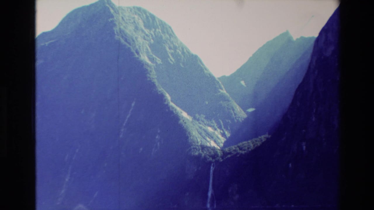 Steep blue-toned mountain valley with a narrow waterfall descending toward a lake or basin.