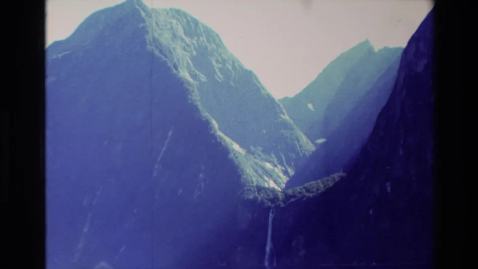 1982 New Zealand 8mm frame showing steep mountain peaks, shadowed ridges, and a thin waterfall in a cool blue film cast.