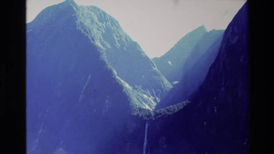 Cool-toned 8mm view of New Zealand mountain slopes, distant peaks, and a slender waterfall descending into the valley.