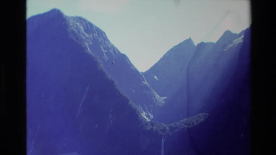 1982 New Zealand home-movie frame with a shifted view across steep alpine slopes and a narrow waterfall below the ridgeline.