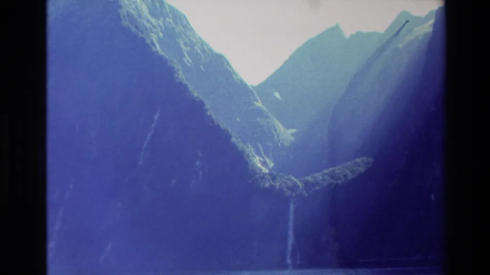 Final 8mm frame of the New Zealand mountain landscape, with jagged peaks, dark valley walls, and a waterfall dropping toward a lake or basin.