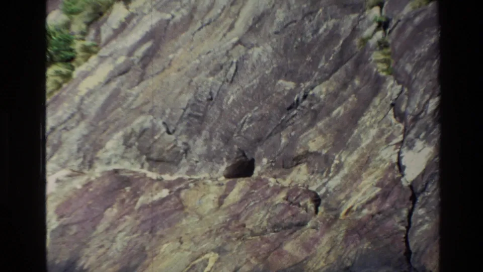1982 New Zealand 8mm frame showing a close rocky cliff surface with cracks, color variation, and a small dark ledge opening.