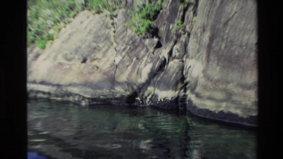 1982 New Zealand 8mm frame of a rock wall meeting calm water, with vegetation above and a shadowed recess near the base.