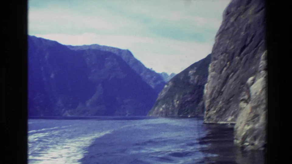 1982 New Zealand 8mm frame looking across blue fjord water toward steep mountains beneath a pale sky.