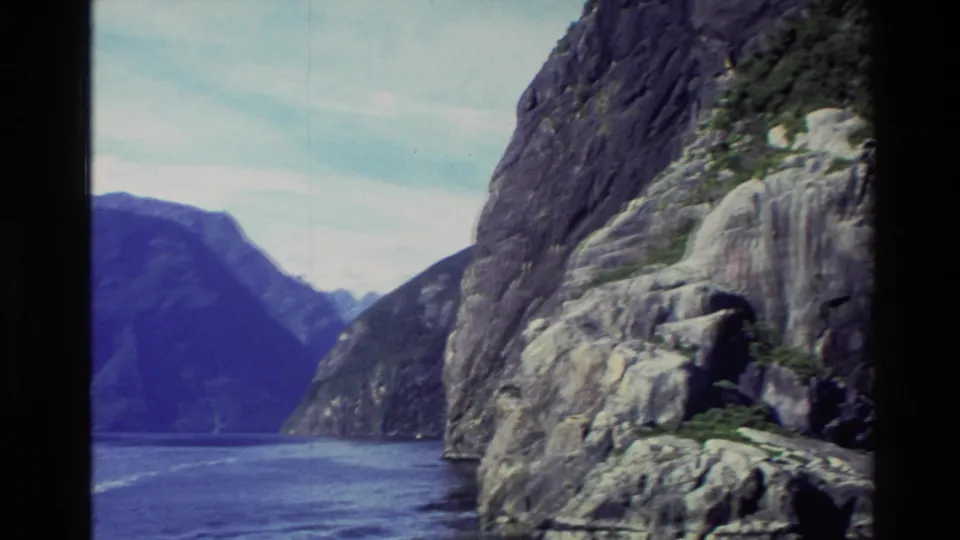 1982 New Zealand 8mm frame showing a close rocky fjord wall, blue water, and distant mountain slopes under cloudy sky.