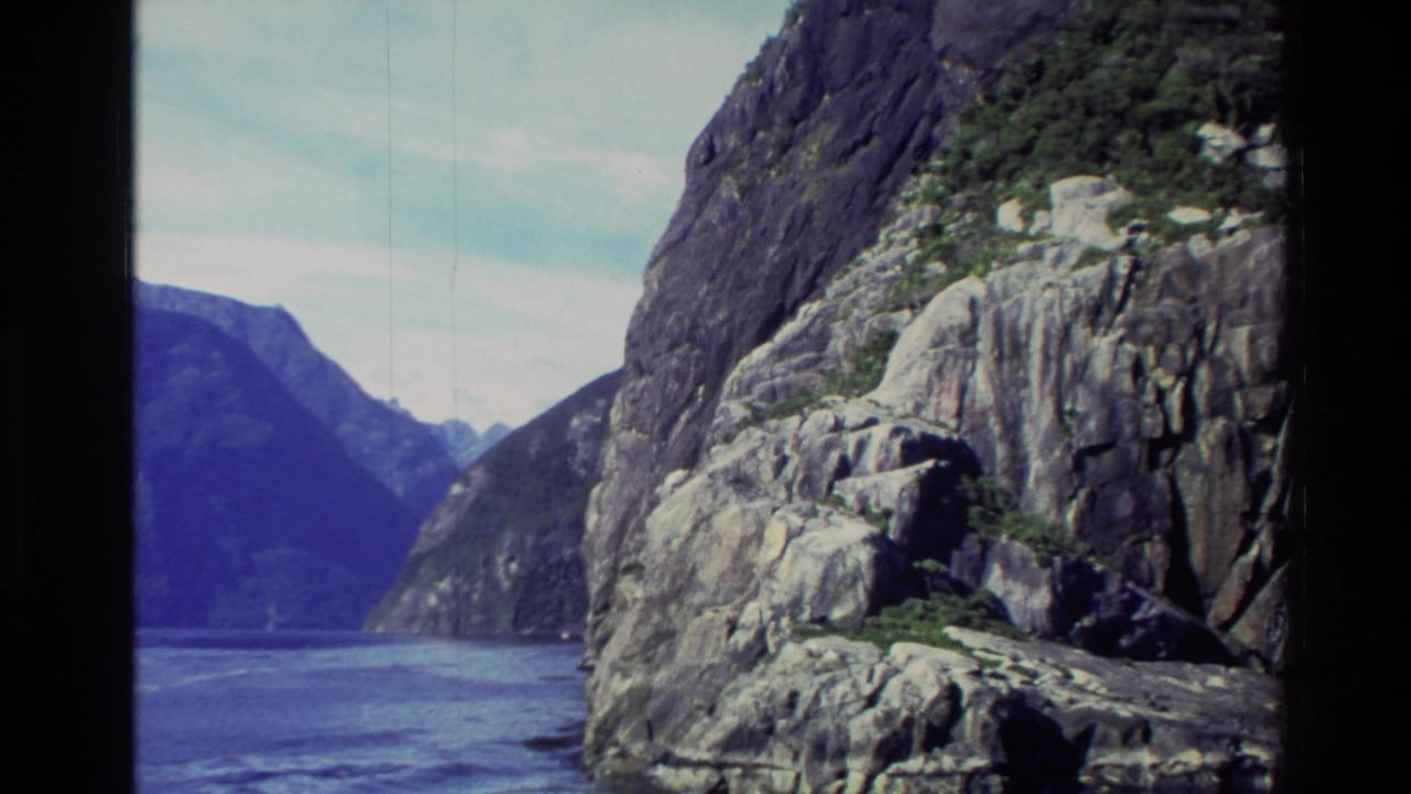 1982 New Zealand 8mm frame showing a boat-view pass by steep coastal rock faces beside deep blue water and distant mountains.