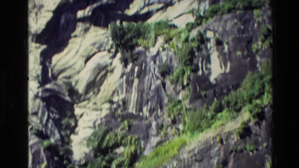 1982 New Zealand 8mm frame showing a close boat-side view of rugged cliff textures and vegetation on the shoreline rock.