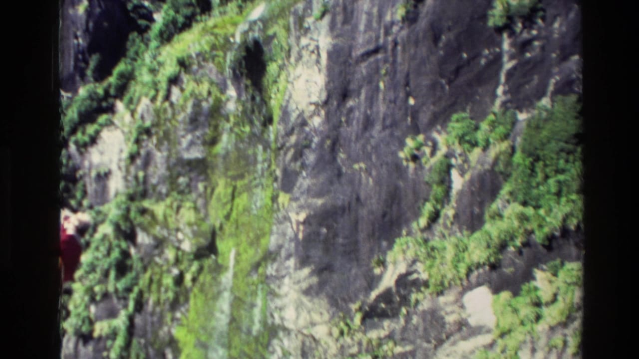 1982 New Zealand 8mm frame showing a moss-covered rock cliff, thin waterfall and partial person in red near the left border.
