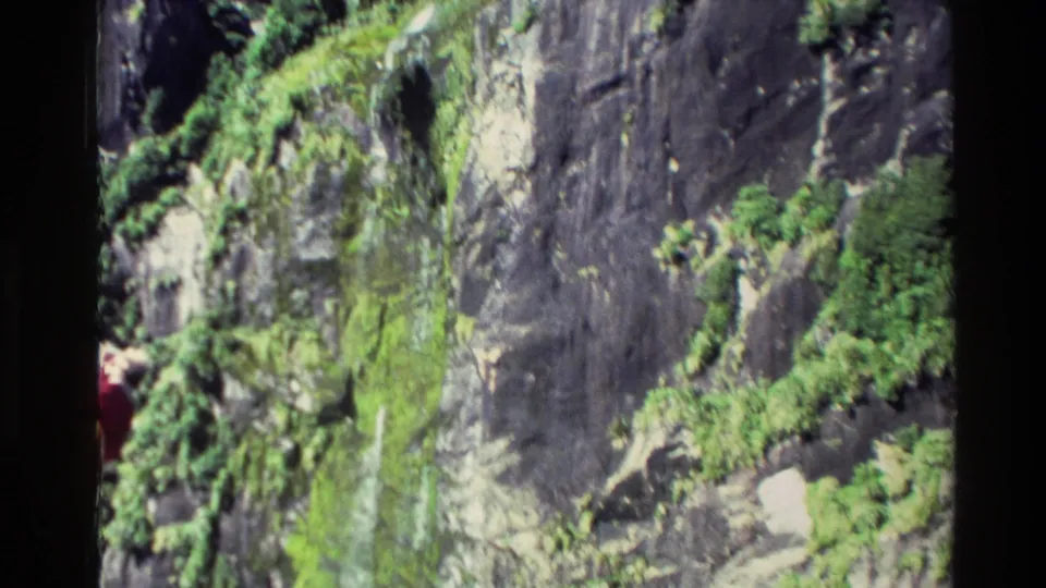 1982 New Zealand 8mm frame showing a moss-covered rock cliff, thin waterfall and partial person in red near the left border.