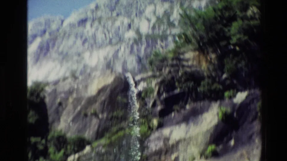 1982 New Zealand 8mm frame with a soft, handheld view of a rocky mountainside, trees and a thin waterfall.