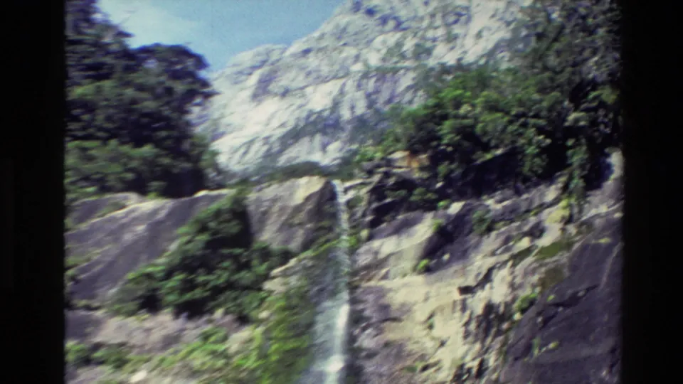 1982 New Zealand 8mm frame showing a wider view of a waterfall, rocky cliff faces, vegetation and pale mountain slopes.