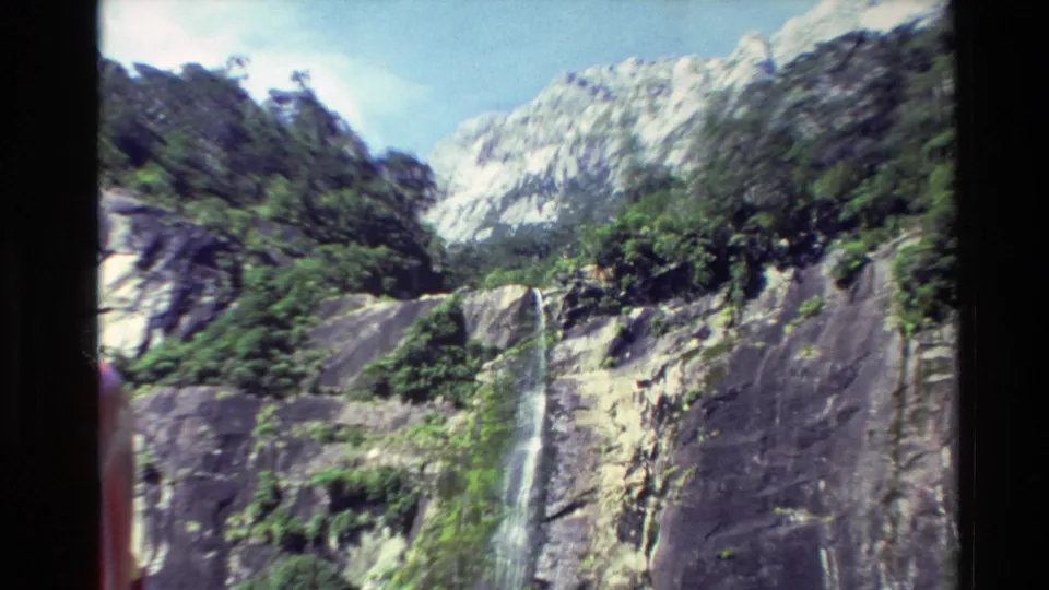 1982 New Zealand 8mm frame showing the waterfall in a wider cliff landscape with vegetation, mountains and a partial red figure at the left edge.