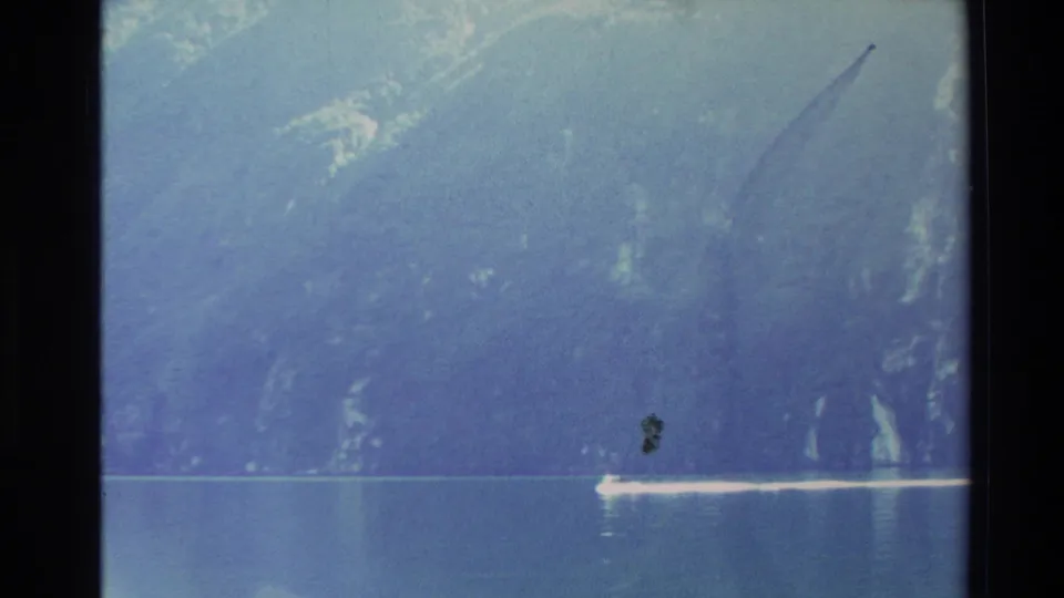 A small boat leaves a pale wake across calm water below hazy New Zealand mountain cliffs in this 1982 home-movie frame.