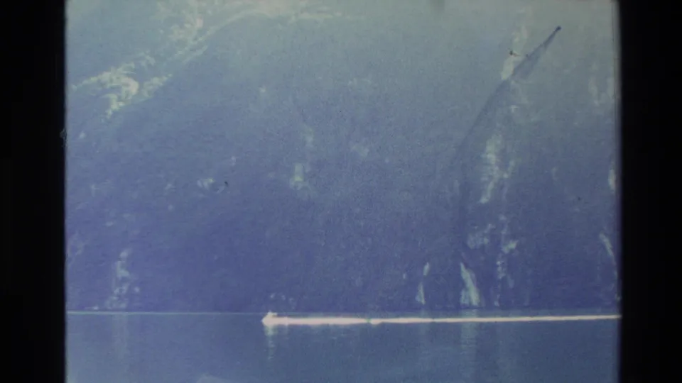 1982 New Zealand 8mm frame showing a boat wake against calm water and steep shadowed cliffs, partially softened by film exposure.