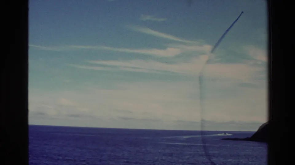 1982 New Zealand coastal view with open water, a distant shoreline, a small boat wake, and dark frame borders.