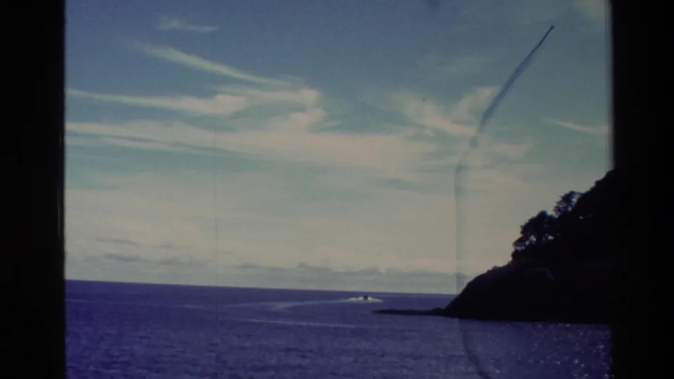 1982 New Zealand 8mm frame with a small boat moving near a dark island edge under streaked clouds.