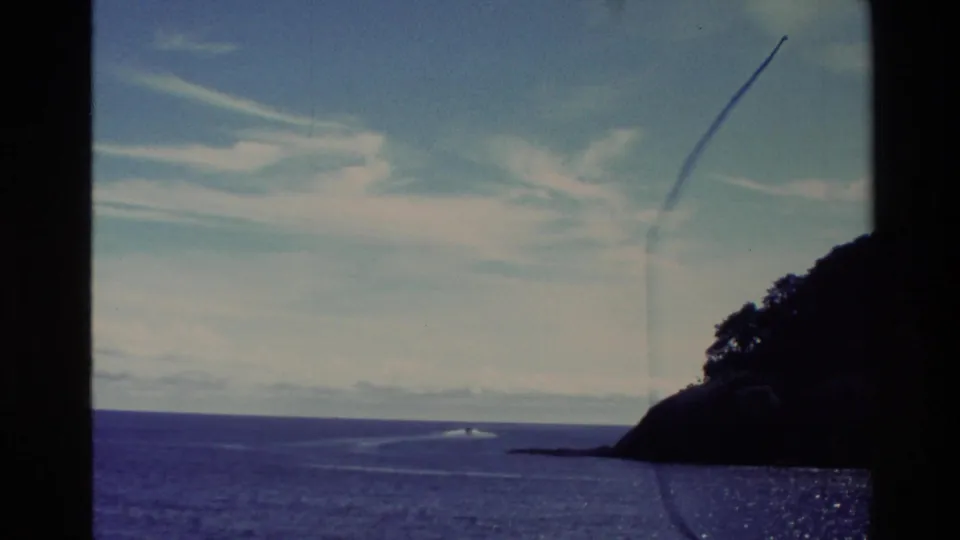 1982 New Zealand coastal home-movie frame showing a distant boat wake, island shoreline, blue water, and visible film marks.