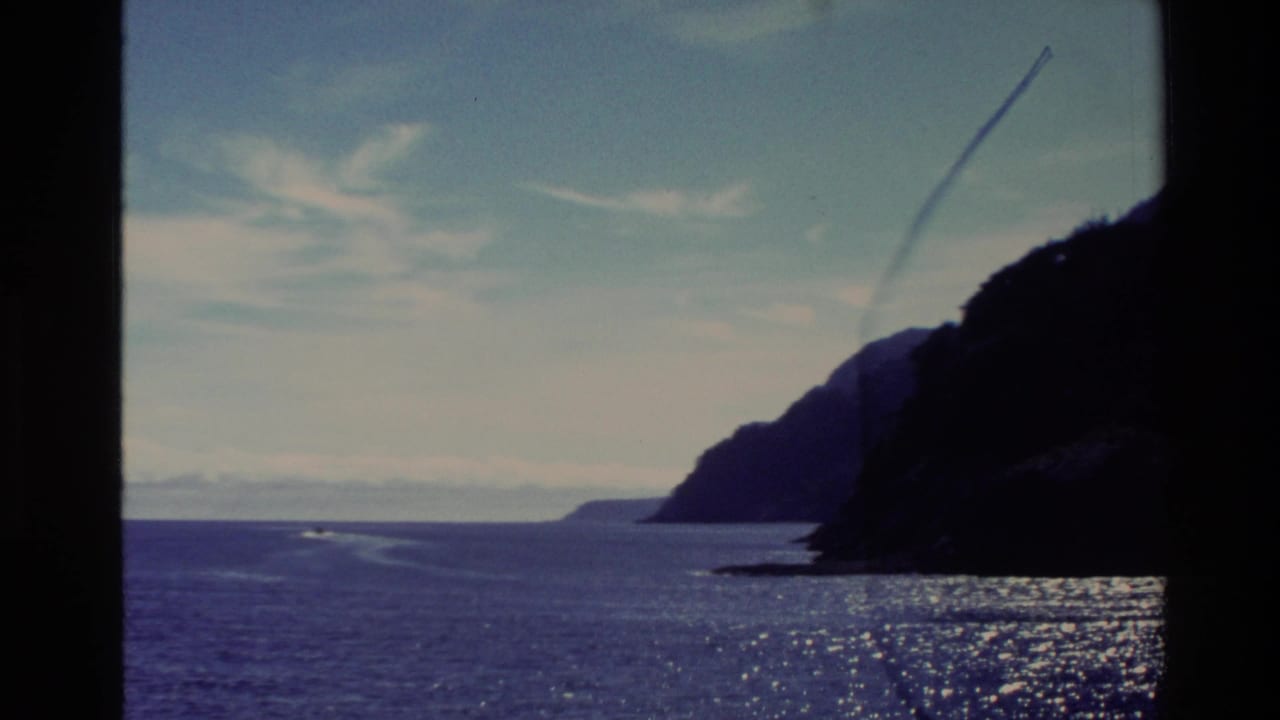 1982 New Zealand 8mm frame showing open ocean, a rugged dark coastline, glinting water, distant wake, and a partly cloudy sky.