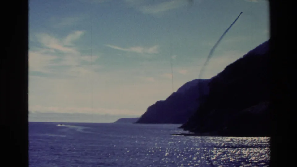 1982 New Zealand coastal view from a moving boat, with a dark headland, shimmering ocean surface, pale clouds, and visible film scratches.