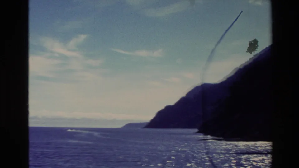 1982 New Zealand 8mm travel frame with open sea, a shadowed mountain coast, distant boat wake, light clouds, and dust on the film.