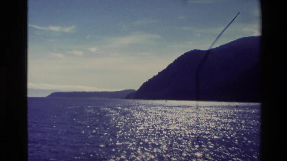 1982 New Zealand boat-travel frame showing a broad glittering ocean, dark mountain slope, distant coastline, and aged 8mm film artifacts.
