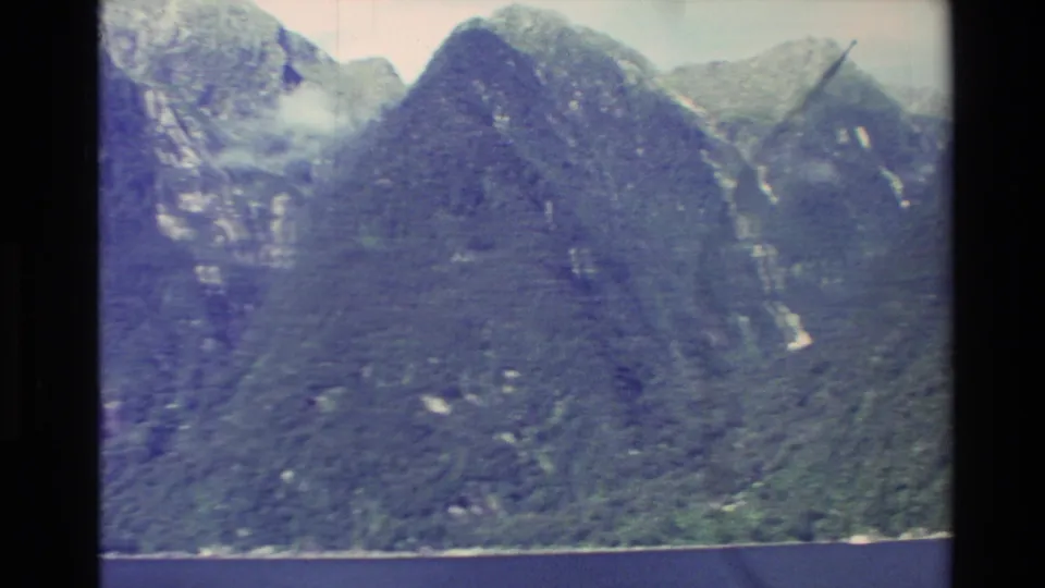1982 New Zealand frame of steep green fjord mountains with rocky faces, snow patches, and a thin shoreline.